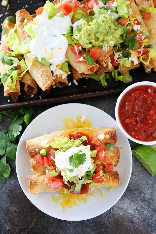 Baked Chicken Taquitos on plate with toppings, a bowl of salsa, and platter of taquitos. 