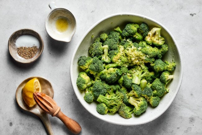 A bowl of broccoli florets next to a bowls with juiced lemon, seasoning, and garlic.
