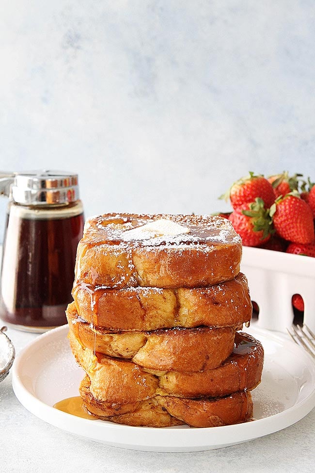 large stack of brioche French toast on plate with butter, powdered sugar, and maple syrup. 