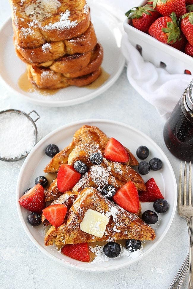 French Toast cut in slices on plate with powdered sugar, butter, syrup, and berries. 