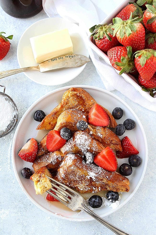 French toast slices on plate with powdered sugar, syrup, berries, and fork. 