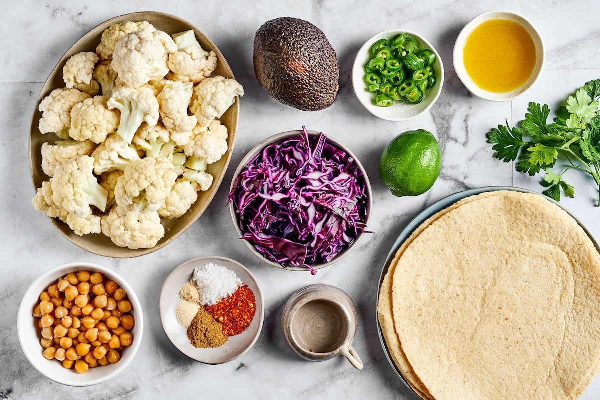 Overhead view of ingredients for roasted cauliflower and chickpea tacos: tortillas, raw cauliflower, car cabbage, jalapeño slices, a lime, an avocado, canned chicpeas, a bowl of spices, a jar of water, a bowl of olive oil, and a sprig of cilantro
