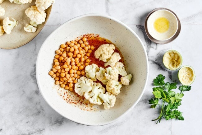 A bowl of marinade with chickpeas and raw cauliflower on top, next to a bowl of cauliflower, squeezed lime halves, cilantro, and a stack of empty bowls