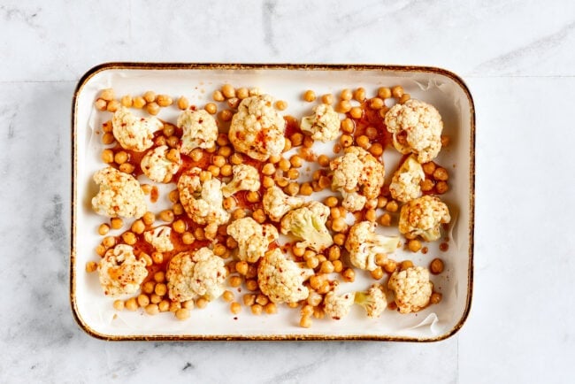 A roasting pan lined with parchment paper and filled with cauliflower and chickpeas that are coated in lime juice, olive oil, chili powder, cumin, onion powder, garlic powder, and salt