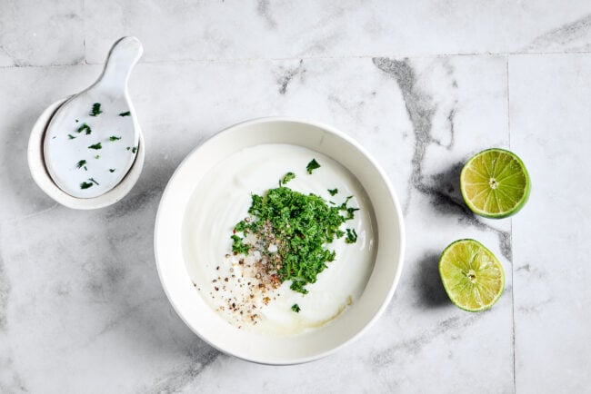 A bowl of greek yogurt with chopped cilantro, salt, pepper, and lime juice on top, next to two lime halves