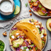 A plate with two roasted cauliflower and chickpea tacos on it, topped with avocado and fresh jalapeño, next to a plate with lime slices and a jar of lime crema, with half an avocado and a plate of tacos next to it