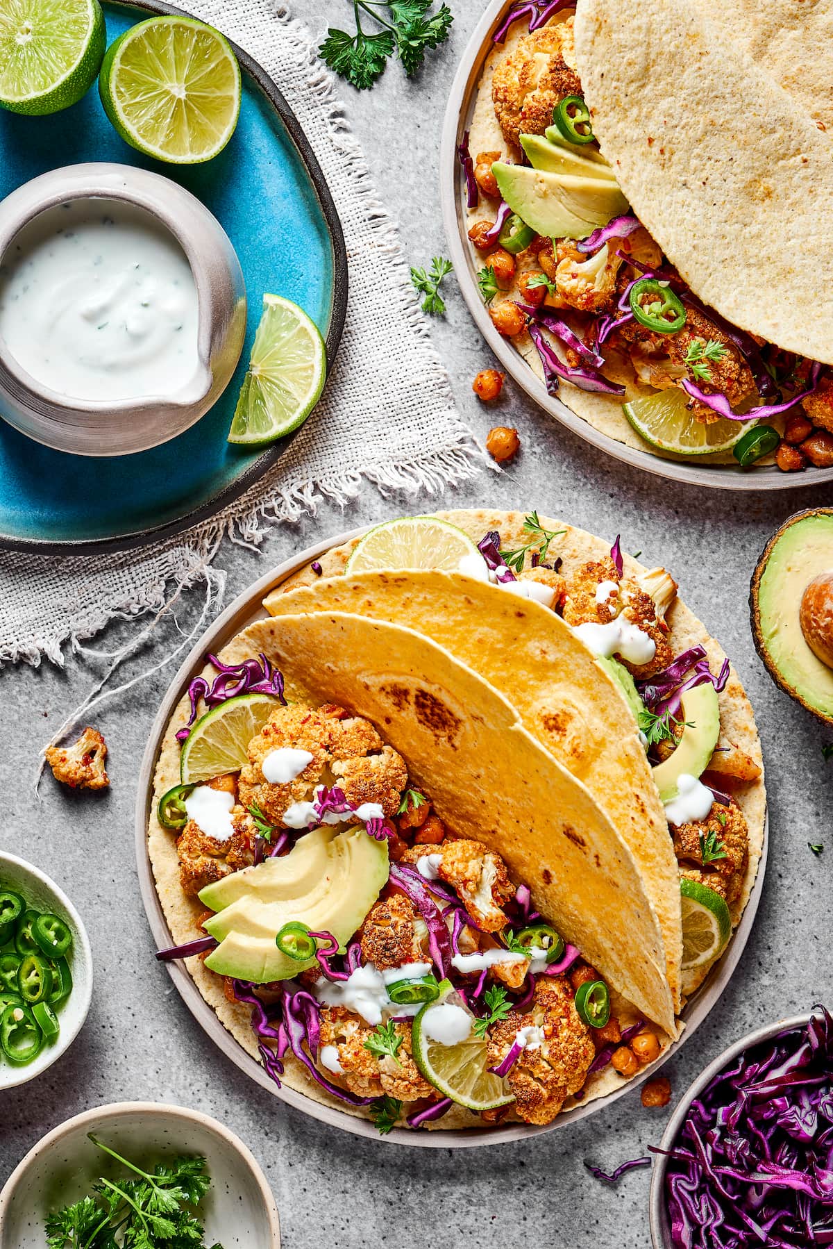 A plate with two roasted cauliflower and chickpea tacos on it, topped with avocado and fresh jalapeño, next to a plate with lime slices and a jar of lime crema, with half an avocado and a plate of tacos next to it