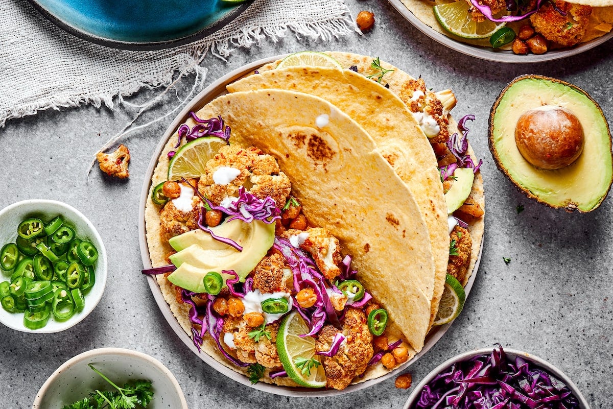 A plate with two roasted cauliflower and chickpea tacos, next to half an avocado and a bowl of jalapeño slices