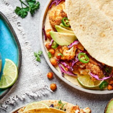 Close up of a roasted cauliflower and chickpea taco on a plate, next to a plate with lime slices and a bowl of lime crema, with a sprig of cilantro