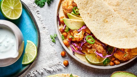 Close up of a roasted cauliflower and chickpea taco on a plate, next to a plate with lime slices and a bowl of lime crema, with a sprig of cilantro