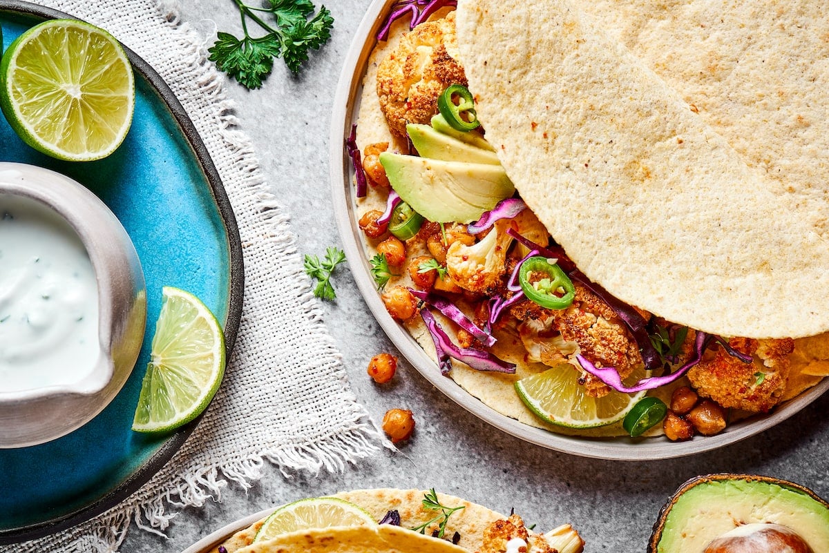 Close up of a roasted cauliflower and chickpea taco on a plate, next to a plate with lime slices and a bowl of lime crema, with a sprig of cilantro
