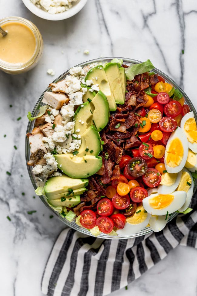 Cobb salad in large glass bowl with jar of dressing on the side and tea towel. 