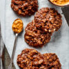 Five no bake cookies on a piece of parchment paper, next to a bowl of peanut butter and a spoonful of peanut butter