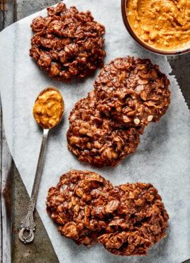 Five no bake cookies on a piece of parchment paper, next to a bowl of peanut butter and a spoonful of peanut butter