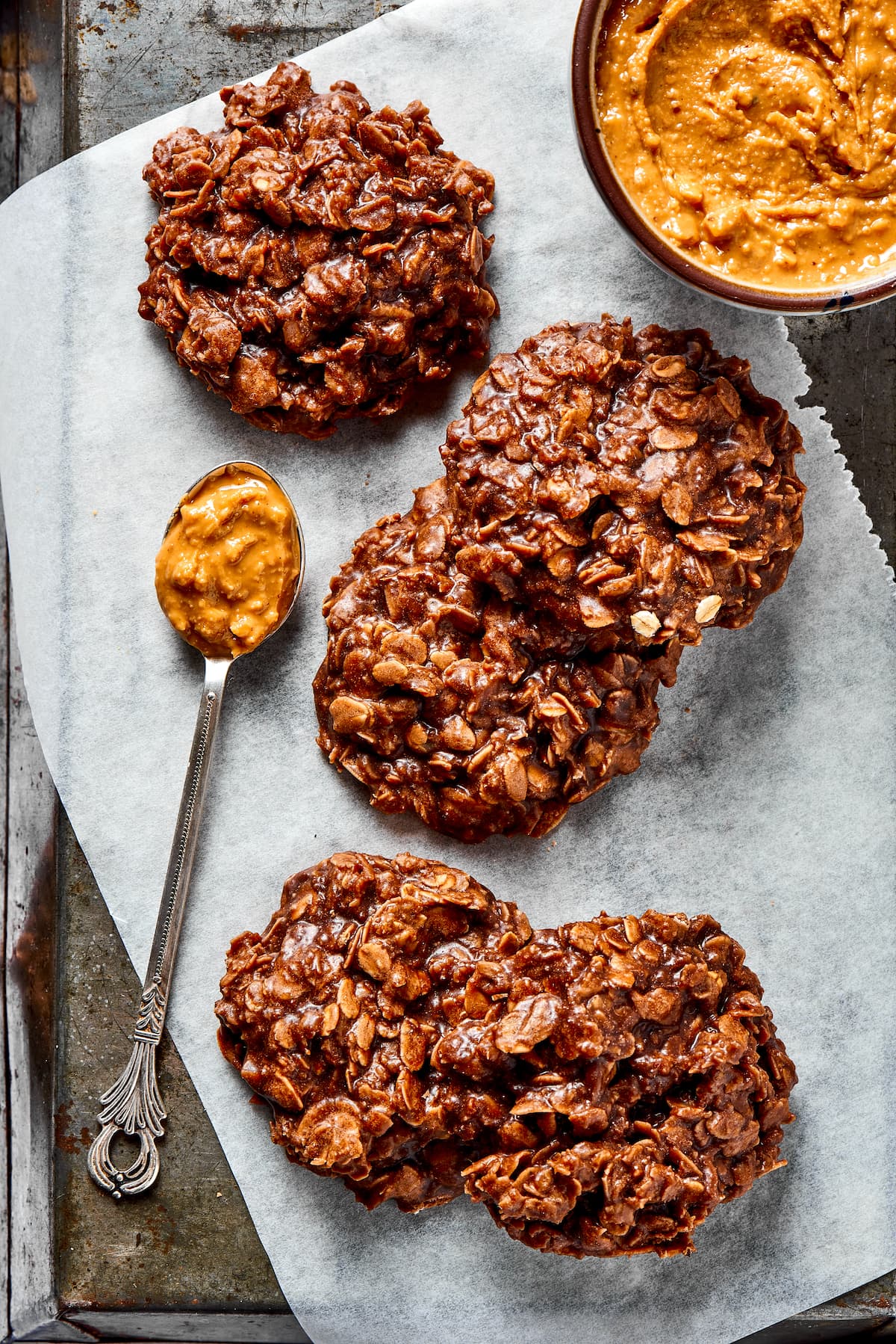 Five no bake cookies on a piece of parchment paper, next to a bowl of peanut butter and a spoonful of peanut butter