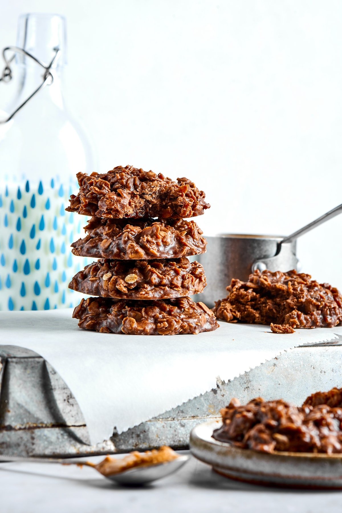 Four no bake cookies stacked on top of each other, with cookies and a saucepan in the background, and a plate of cookies and a spoonful of peanut butter in the foreground