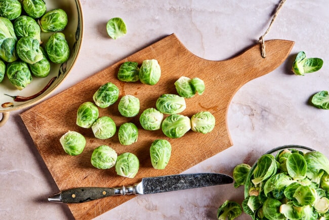 Raw Brussels sprouts on a cutting board, with a knife on the cutting board, next to a bowl of Brussels sprouts and a bowl of the outer layers.