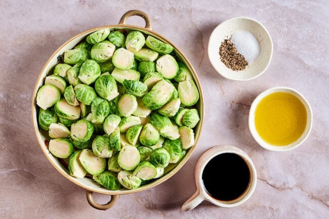A mixing bowl full of raw Brussels sprouts, next to a small container of balsamic vinegar, a small bowl of olive oil, and a small bowl of salt and pepper