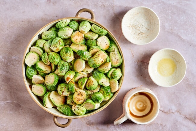 A mixing bowl full of Brussels sprouts, coated in salt, pepper, olive oil, and balsamic vinegar, next to three small empty bowls