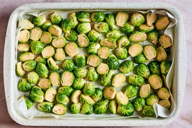 A baking tray with a layer of parchment paper, full of uncooked, seasoned Brussels sprouts