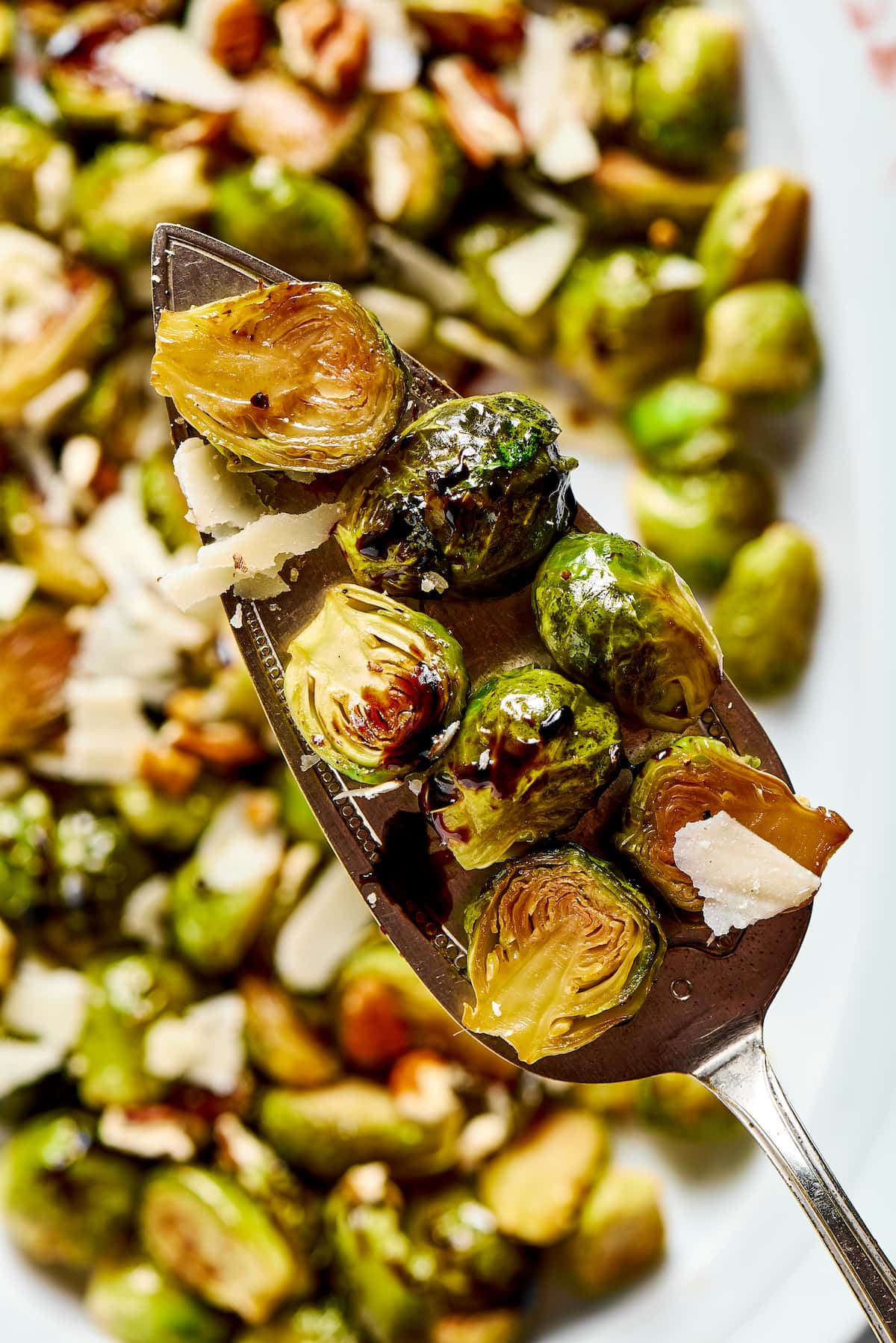 A serving spoon with a serving of roasted Brussels sprouts on it, with a pan of Brussels sprouts covered in parmesan flakes in the background