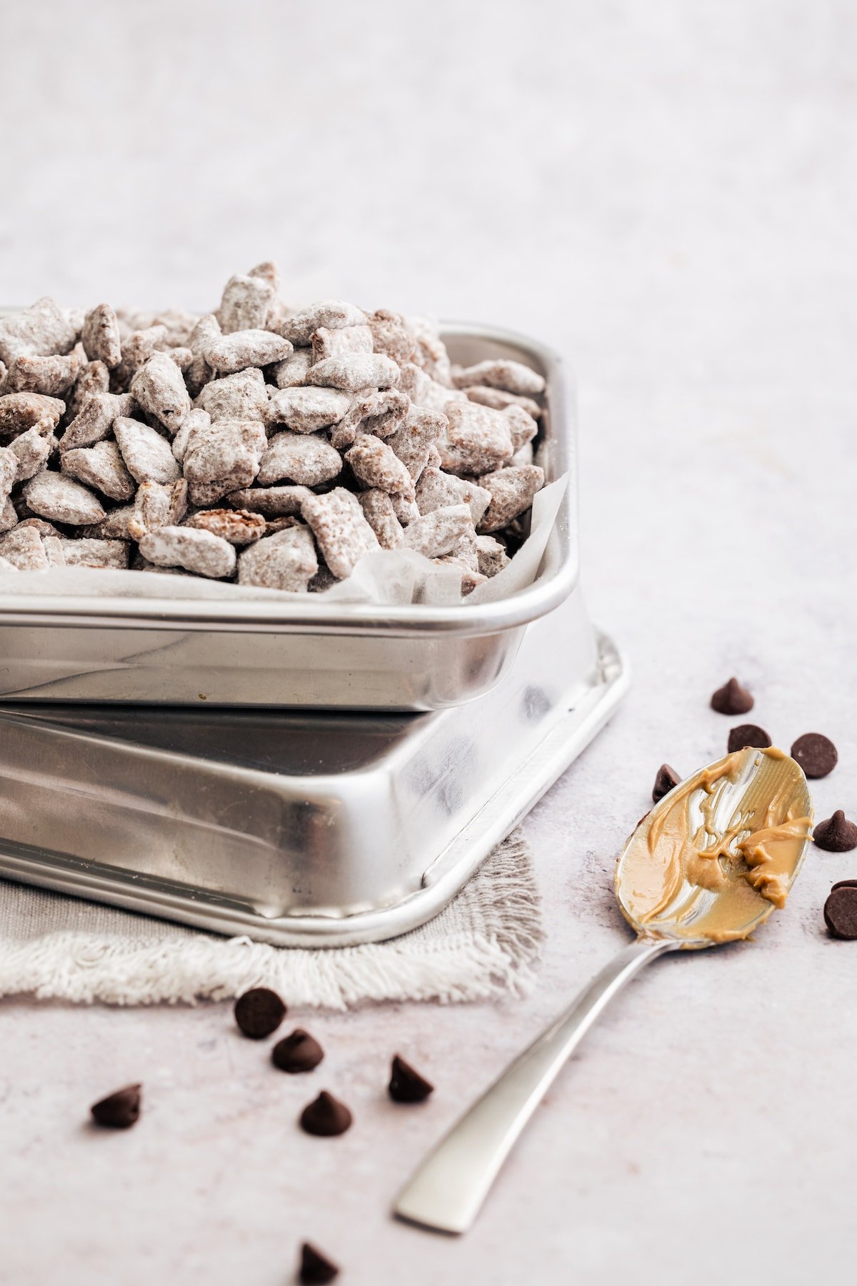 Puppy chow in a parchment-lined metal pan, stacked on top of another pan next to a spoon covered with peanut butter.