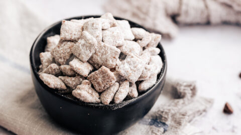 A bowl of puppy chow on a grey cloth, surrounded by scattered puppy chow and a spoon covered with peanut butter.