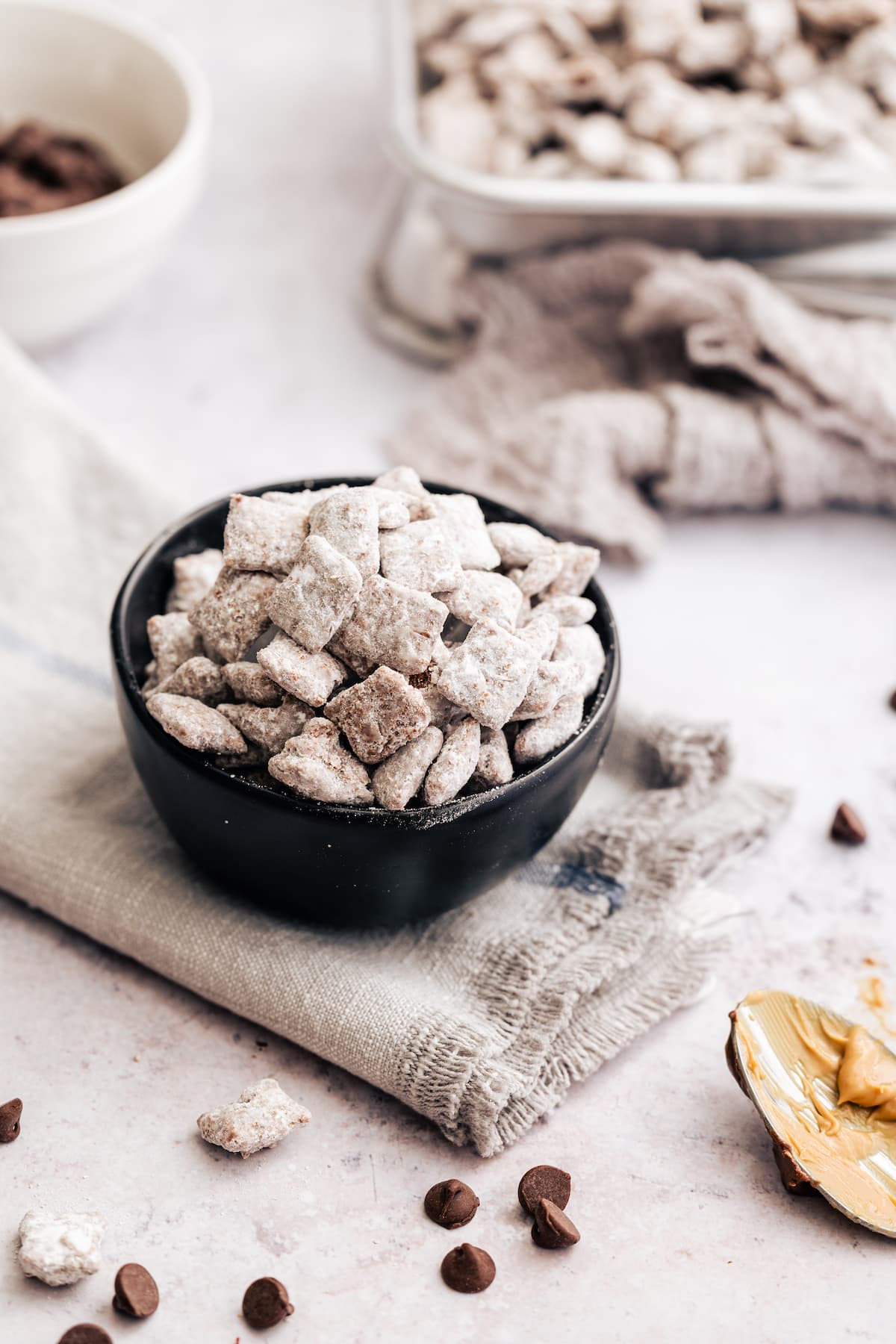 A bowl of puppy chow on a grey cloth, surrounded by scattered puppy chow and a spoon covered with peanut butter.