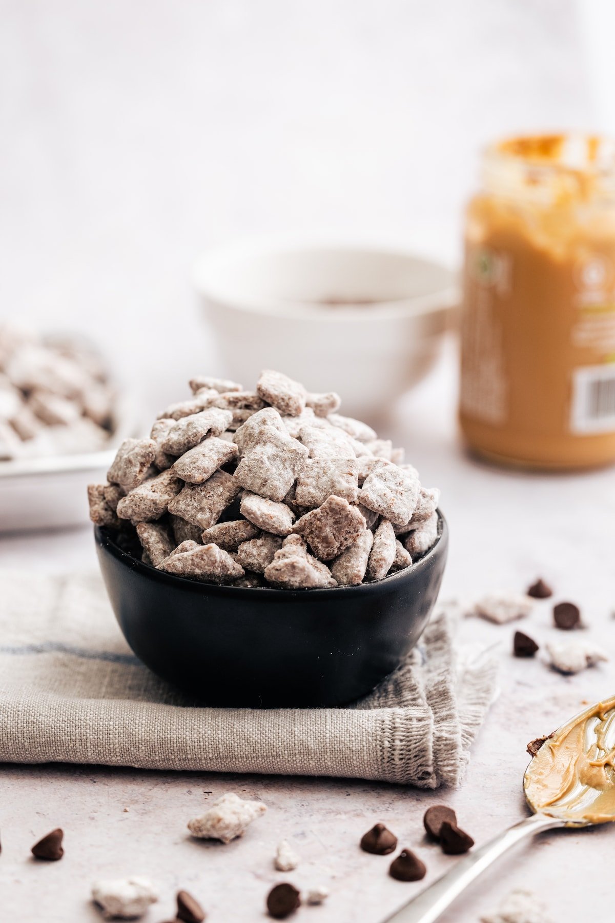 A bowl of puppy chow on a grey cloth with a jar of peanut butter in the background.