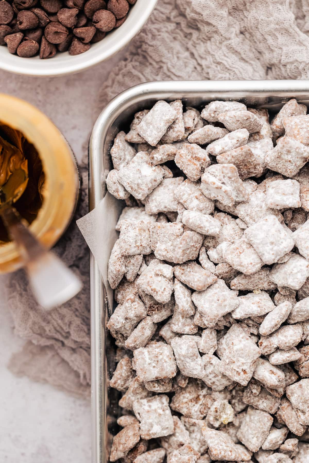 Overhead view of the corner of a metal pan filled with puppy chow next to a jar of peanut butter and a bowl of chocolate chips.