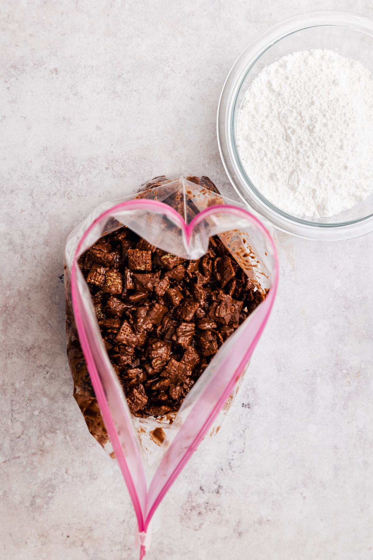 Overhead view of chocolate and peanut butter-coated Chex cereal in a large ziploc bag, next to a small bowl of powdered sugar.