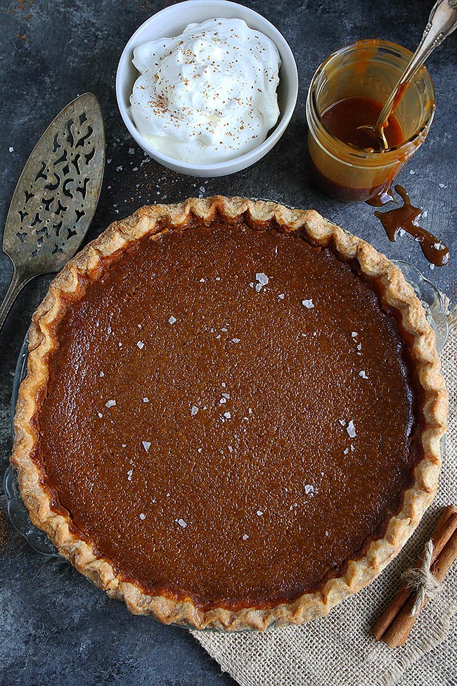 salted caramel pumpkin pie in pie plate with a bowl of whipped cream and jar of salted caramel sauce. 