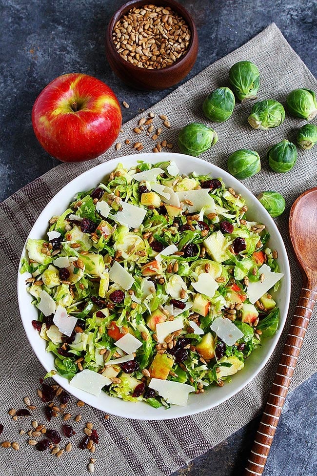 A bowl of shaved Brussels sprouts salad topped with shaved parmesan, on top of a place mat, next to a whole apple, nine whole Brussels sprouts, a wooden serving spoon, a bowl of sunflower seeds, and scattered sunflower seeds and dried cranberries. 