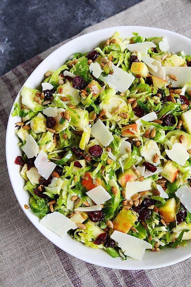 This raw shaved Brussels sprouts salad is crisp and refreshing, with apples and a maple dressing. It's perfect for Thanksgiving or Christmas! A bowl of shaved Brussels sprouts salad, with large chunks of apple and dried cranberries, topped with shaved parmesan.