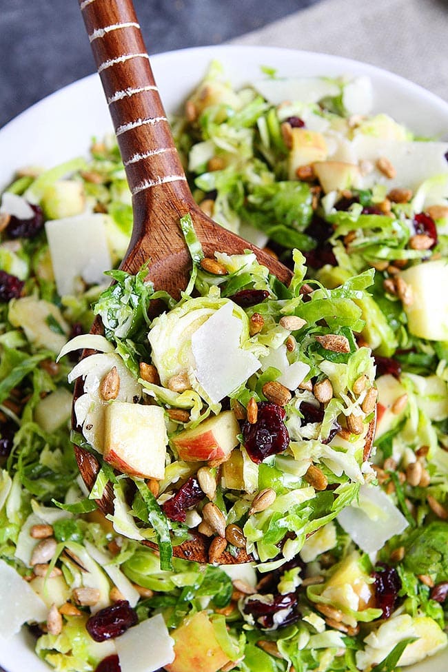 Overhead view of a wooden serving spoon full of shaved Brussels sprouts salad, with slices of apples, dried cranberries, sunflower seeds, and shaved parmesan cheese, with a bowl of the salad in the background. 