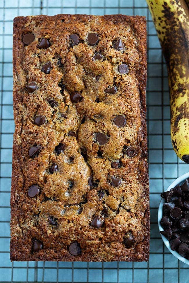 Overhead view of a loaf of chocolate chip banana bread on a wire cooling rack, next to an overly ripe banana and a bowl of chocolate chips.