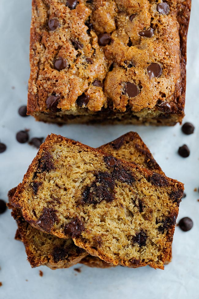 Half a loaf of chocolate chip banana bread next to a stack of three pieces of banana bread, with chocolate chips surrounding them.