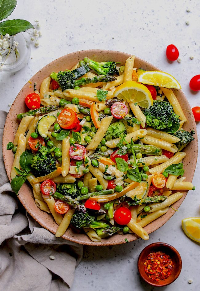 Pasta Primavera in large bowl with fresh herbs and lemon wedges.