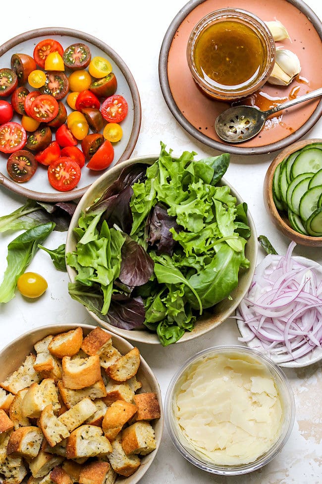 mixed greens in bowl, grape tomatoes on plate, balsamic dressing, red onion slices in small bowl, homemade croutons in bowl, and shaved parmesan cheese in small bowl. 