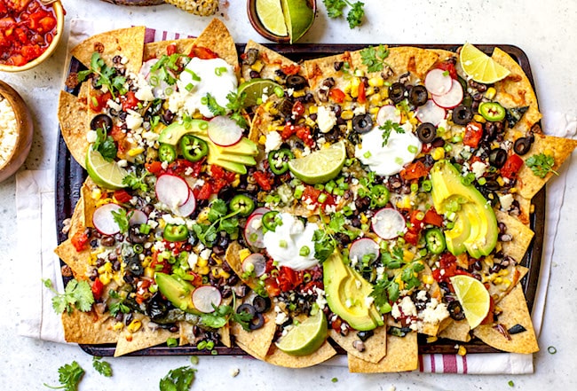 A tray of vibrant, loaded nachos topped with avocado slices, lime wedges, chopped tomatoes, black olives, jalape&ntilde;o slices, sour cream, corn, cilantro, and cheese crumbles. Two small bowls with extra toppings are placed nearby on a striped cloth.