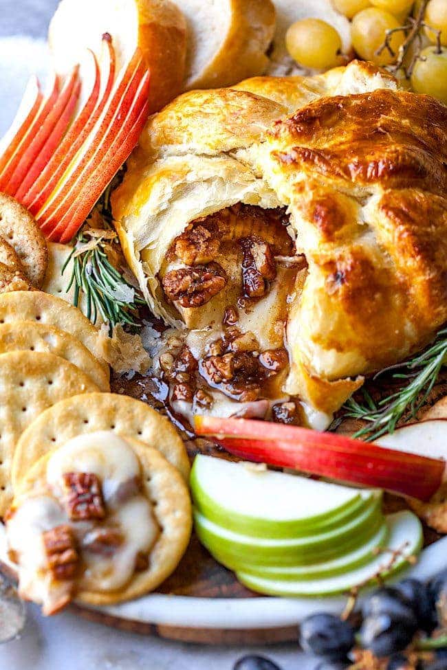 Baked Brie in puff pastry with pecans on platter with crackers, apple slices, and baguette slices. 