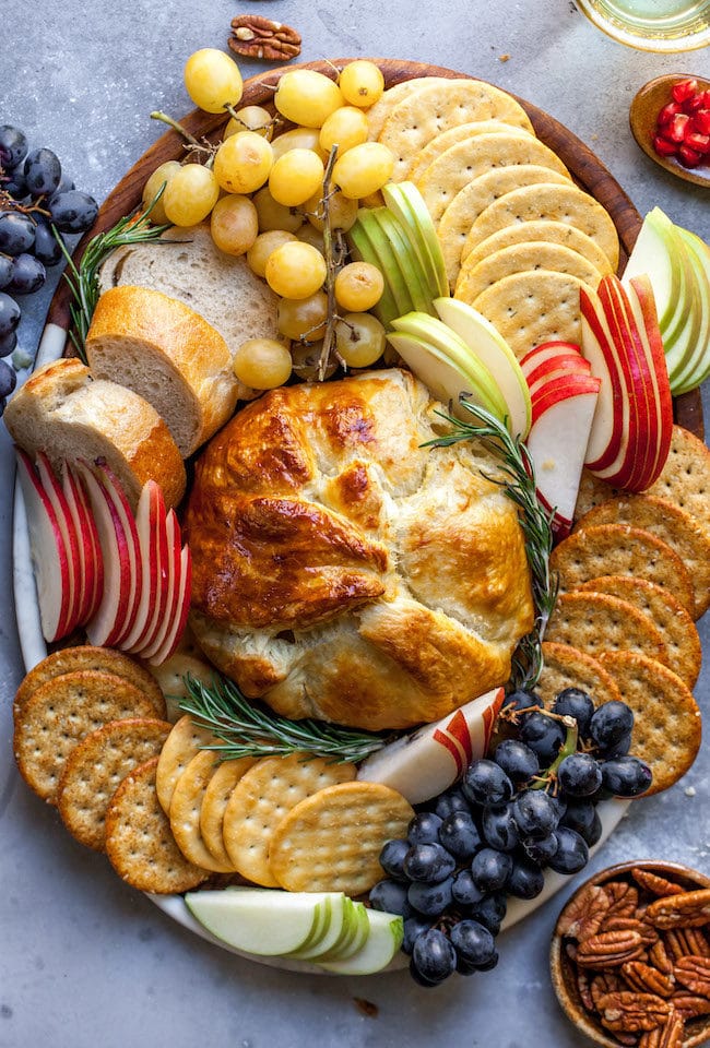 Baked Brie in puff pastry on platter with crackers, fruit, baguette slices, and herbs.