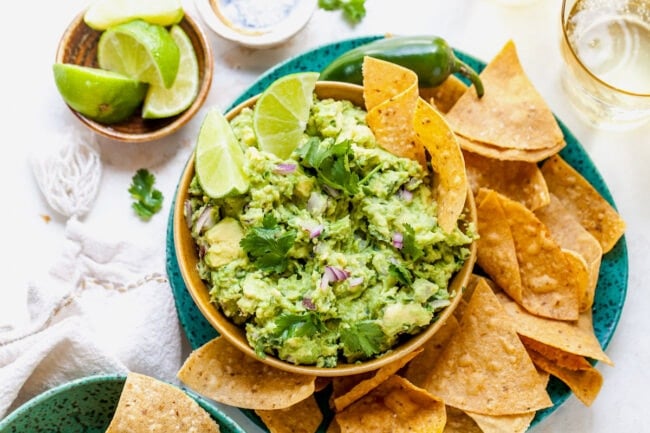 Guacamole Recipe in bowl with corn tortilla chips on plate with a small bowl of lime wedges. 
