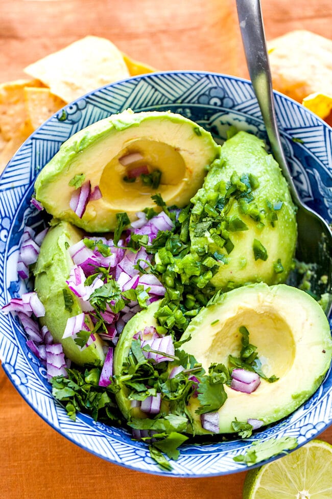 avocados in bowl with spoon with red onion, jalapeño, and cilantro.