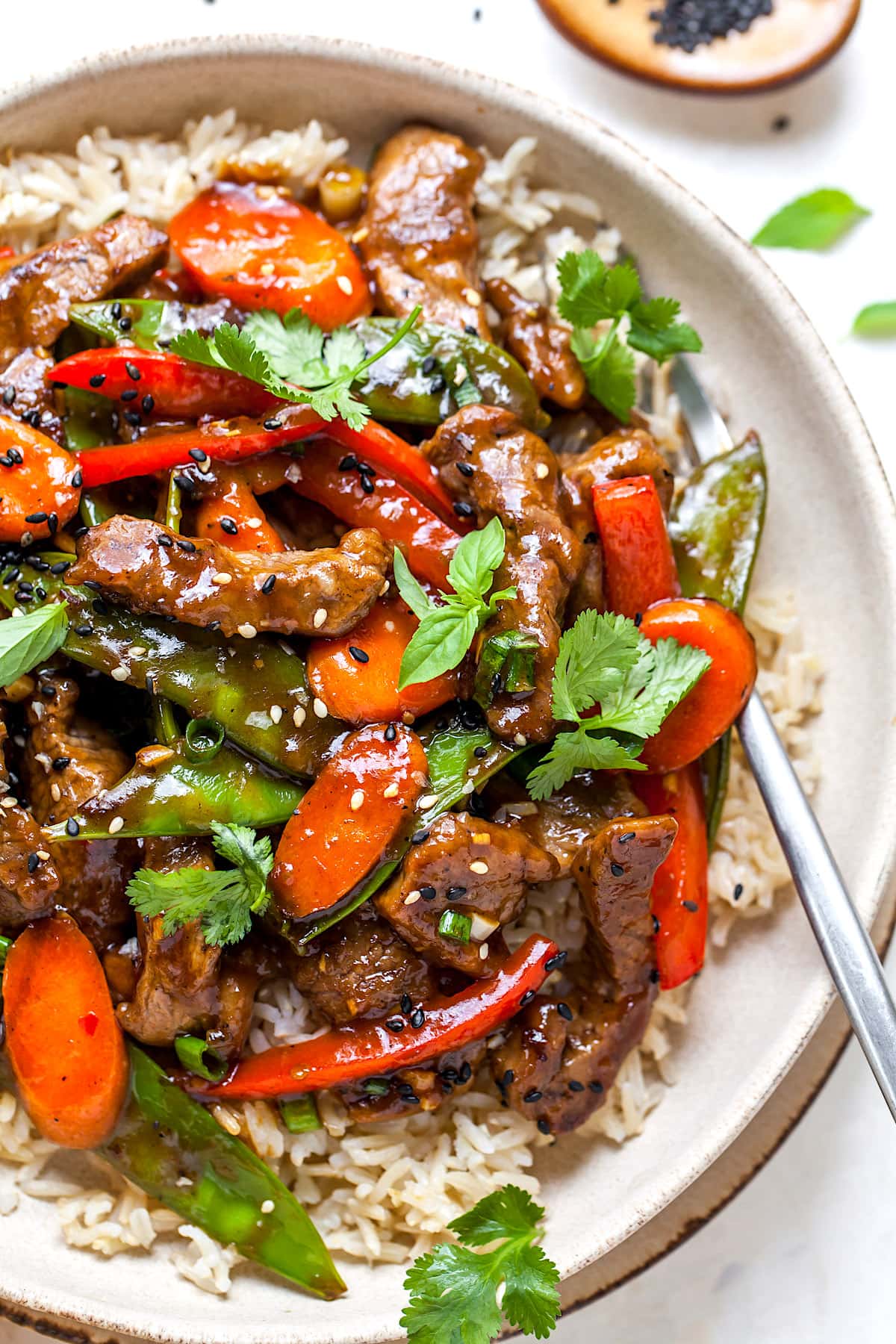 close up of beef stir fry with vegetables and herbs in bowl with rice and fork.