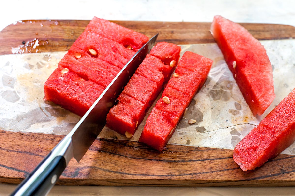 Watermelon being cut into cubes with a knife on wood cutting board. 