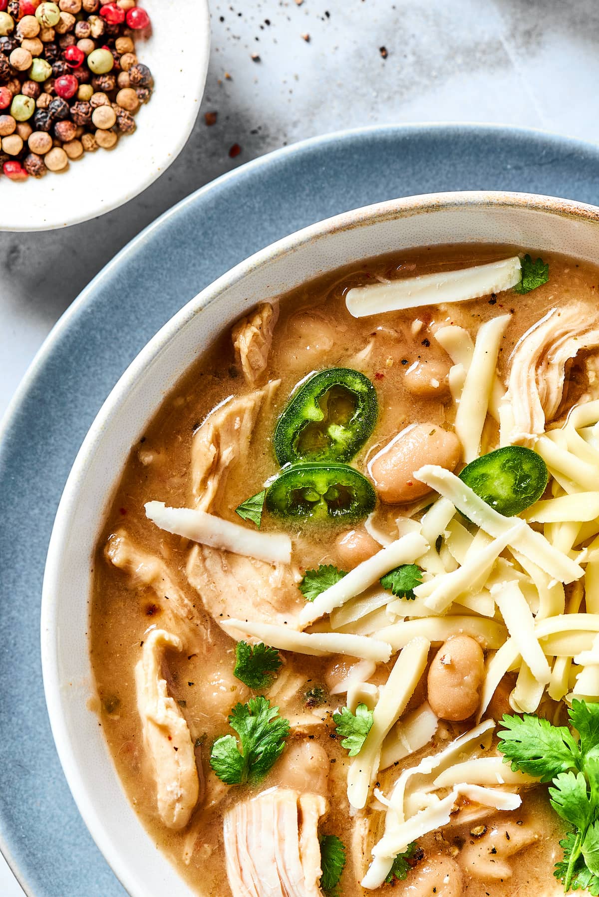 Close up overhead view of a bowl of white chicken chili garnished with fresh cilantro, next to a small bowl of rainbow peppercorns.