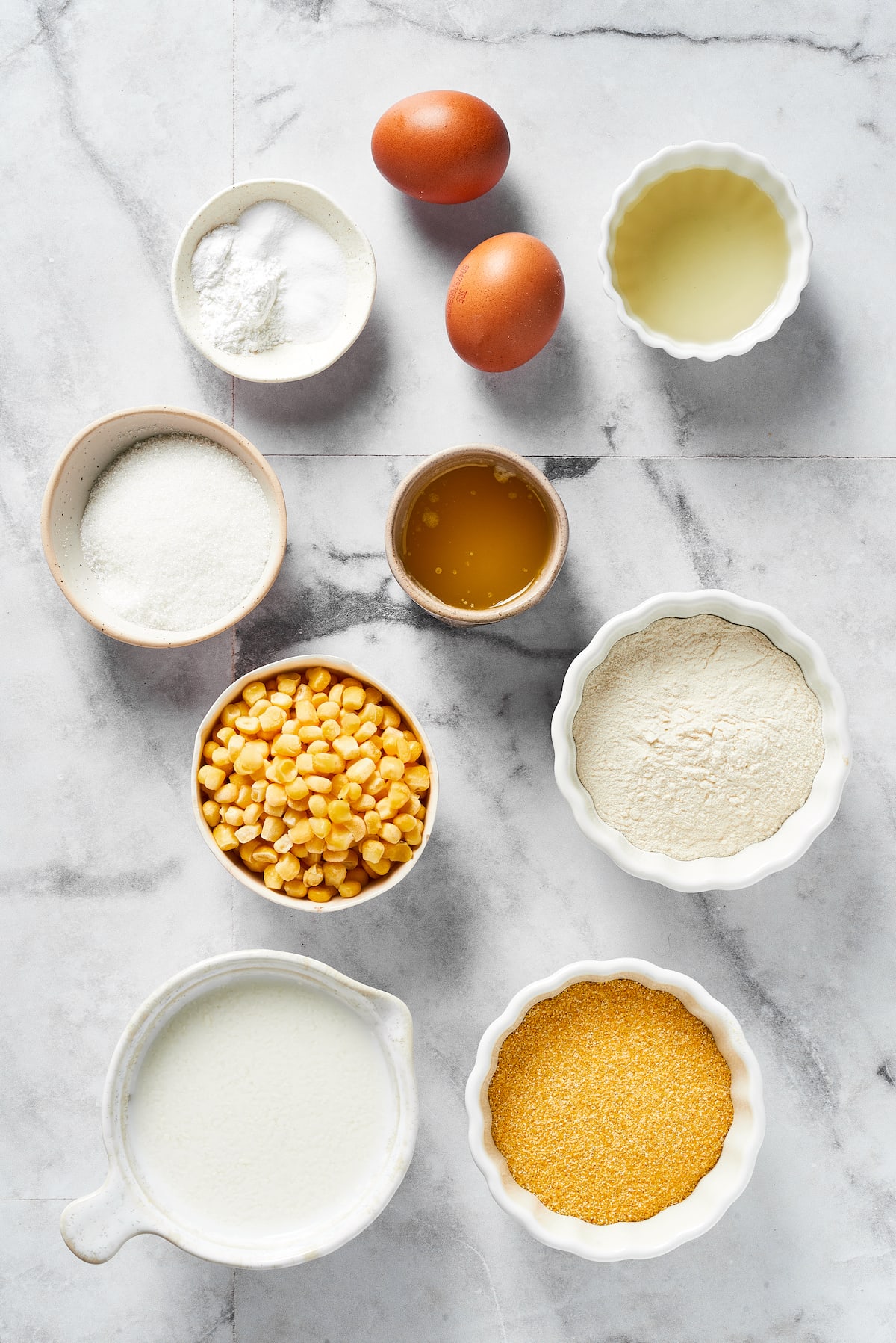 Overhead view of the ingredients needed for cornbread: a bowl of cornmeal, a bowl of flour, a bowl of buttermilk, a bowl of corn, a bowl of melted butter, a bowl of oil, a bowl of sugar, a bowl of salt, baking soda, and baking powder, and eggs