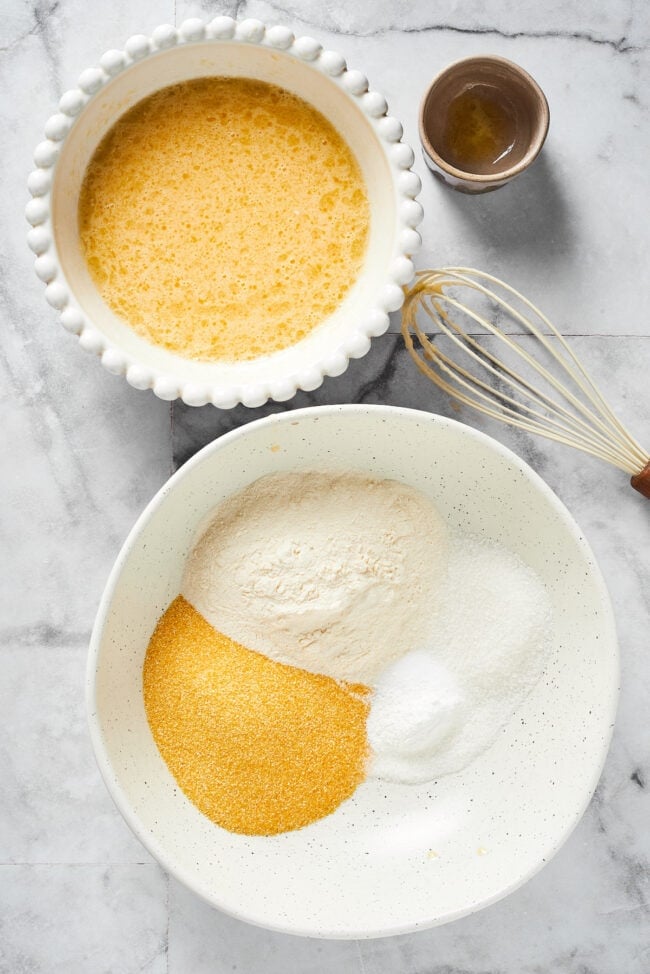 A bowl with cornmeal, flour, salt, sugar, baking soda, and baking powder, unmixed, next to a bowl with eggs, buttermilk, melted butter, and oil, mixed together, next to a whisk