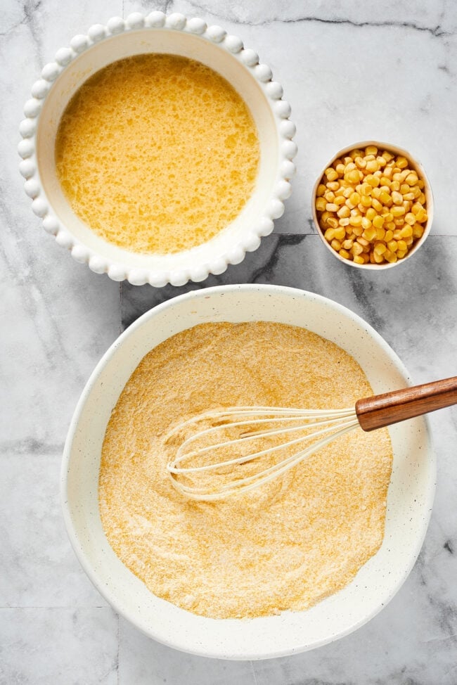 A bowl with cornmeal, flour, salt, sugar, baking soda, and baking powder, mixed together with a whisk in the bowl, next to a bowl with eggs, buttermilk, melted butter, and oil, mixed together, next to a bowl of corn